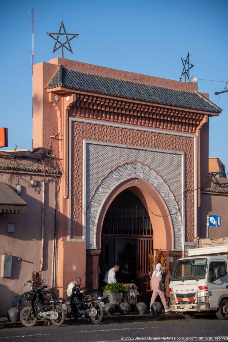 Large horseshoe-arched entrance with carved geometric surround, wooden gate, motorcycles, and a small vehicle against pink walls.