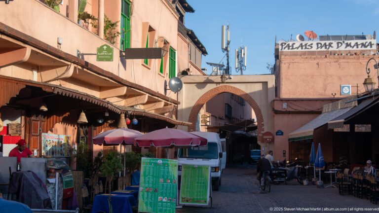 Narrow Marrakech lane with cafés, awnings, scooters, delivery vehicles, brick arch, and Palais Dar D'Mana signage.