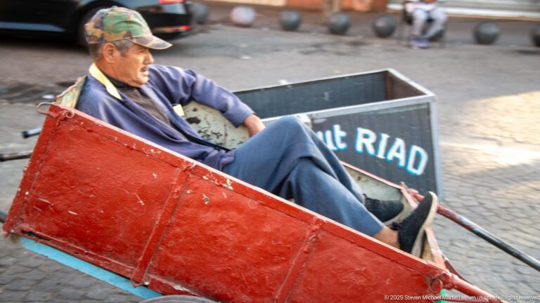 Man seated inside a worn red two-wheeled handcart with another metal cart behind him on a paved street.