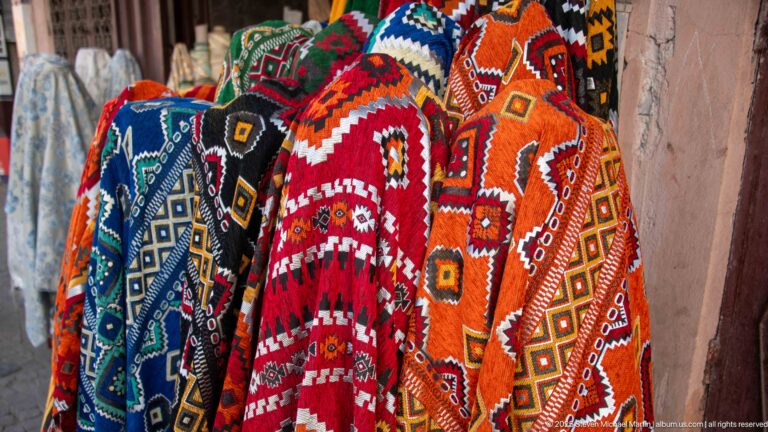 Bright red, orange, blue, and black patterned textiles hanging in a dense vertical display against a pink wall.