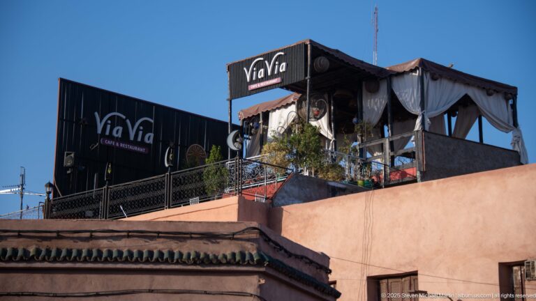 ViaVia Café & Restaurant rooftop terrace with black signs, shaded seating, potted plants, and pink buildings below.