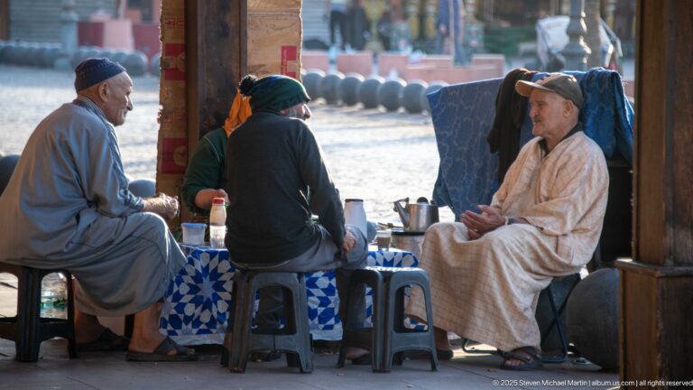 Three men seated on low stools around a small outdoor table with a metal teapot and glasses in Marrakech.