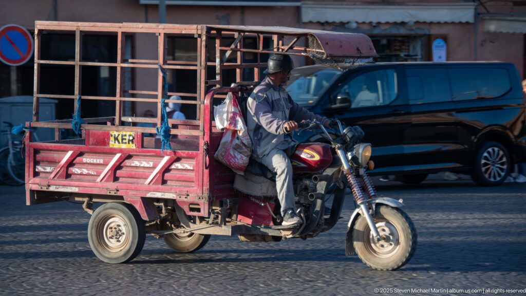Driver riding a red three-wheeled motorcycle cargo vehicle with open metal rack beside a black car in Marrakech.