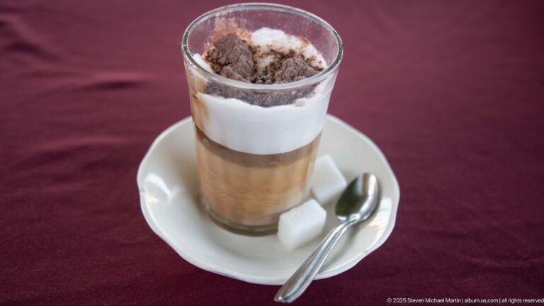 Tall layered coffee in a glass with white foam and cocoa, served on a saucer with spoon and sugar cubes.