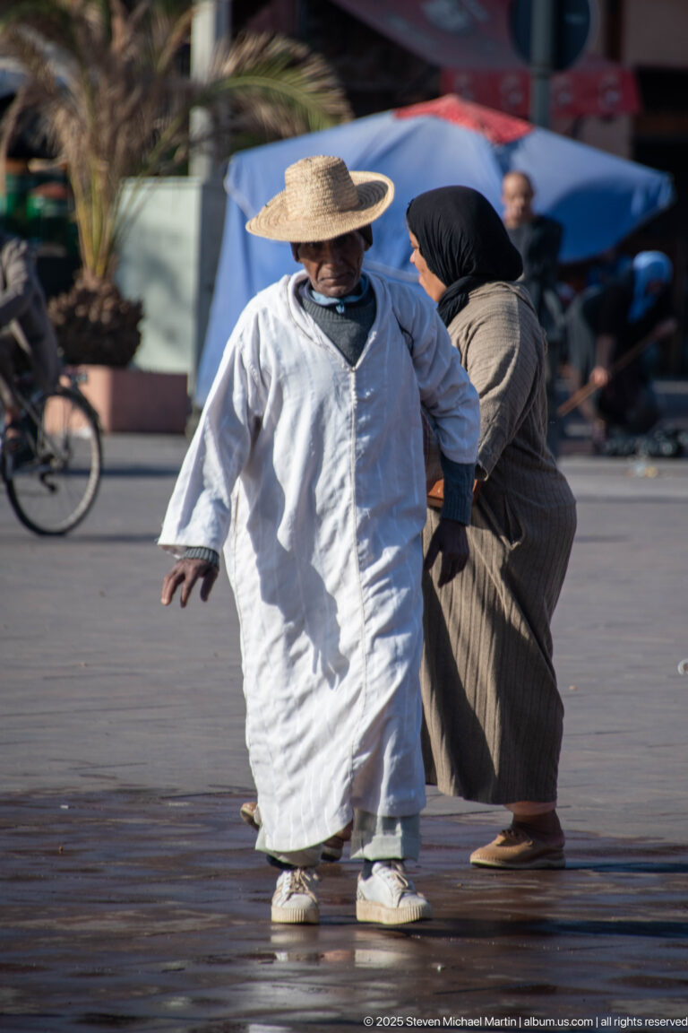 Two women walking across a Marrakech square, one in a broad straw hat, with bicycles and pedestrians blurred behind.
