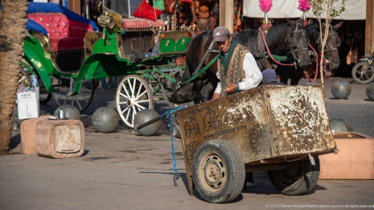 Man pulling a large worn handcart with green horse-drawn carriages and harnessed horses waiting in the background.