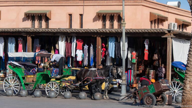 Green horse-drawn carriages beside a clothing shop with hanging garments and spherical bollards along the street edge.