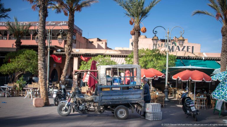 Café Mazel frontage with palm trees, umbrellas, outdoor tables, scooters, and a three-wheeled cargo motorcycle in front.