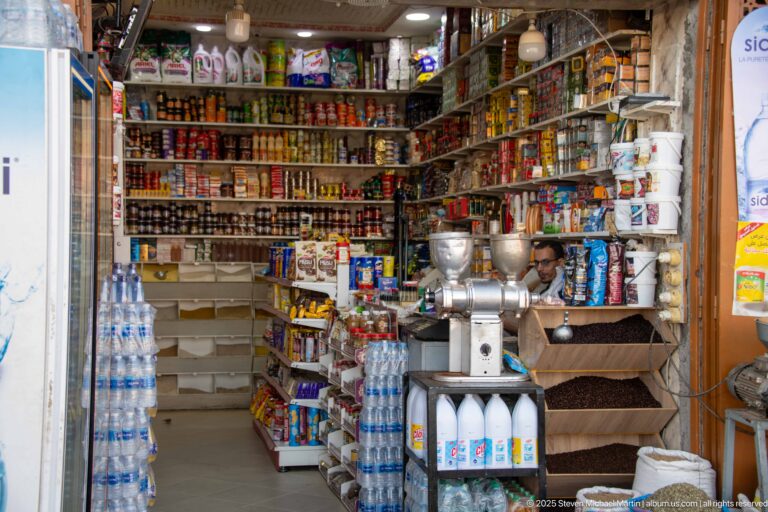 Small Marrakech grocery packed with shelves of tins, jars, bottles, boxes, water, and two metal grinders at the entrance.