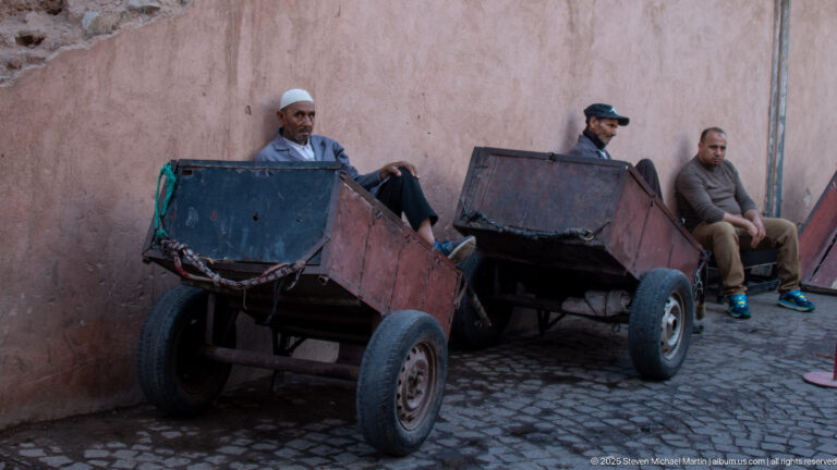 Two large empty handcarts parked against a pink wall with three men resting beside them on a cobbled lane.