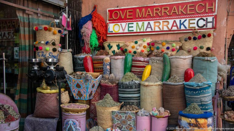 Herbal and spice display with sacks, jars, colorful objects, pom-pom trim, and two signs reading LOVE MARRAKECH.
