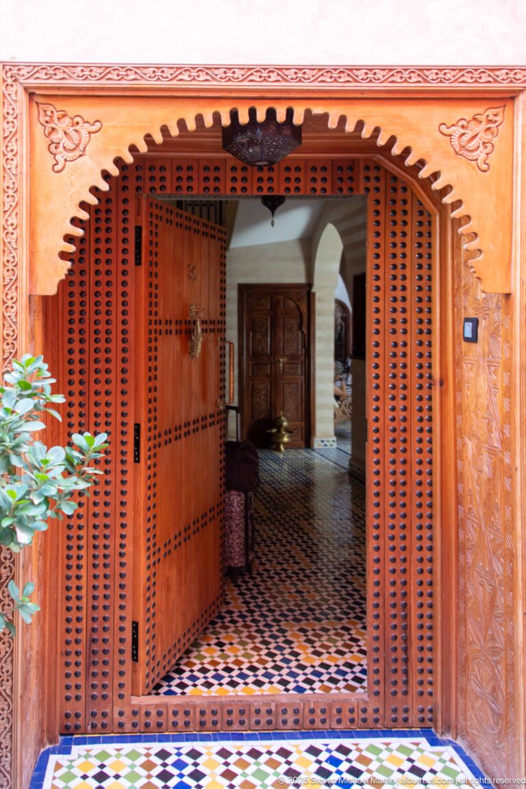Perforated wooden screen and doors framing a tiled passage with white arches and a dark wooden door beyond.
