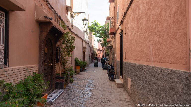 Narrow cobbled Marrakech alley between pink walls with carved doorways, potted plants, pedestrians, and parked motorcycles.