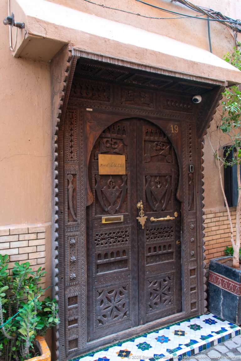 Dark carved wooden double door numbered 19 beneath a canopy, with metal fittings, tiled threshold, and built-in bench.