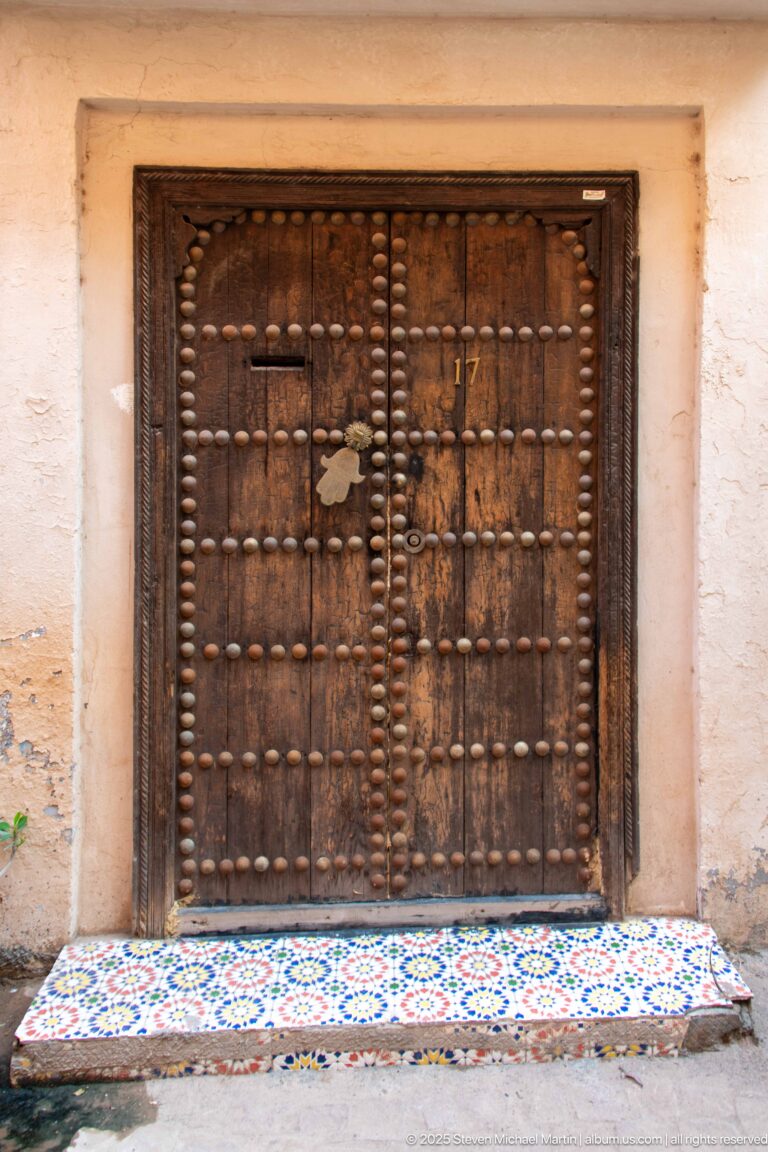 Weathered double wooden door covered with rows of metal bosses above a colorful geometric mosaic-tiled threshold.