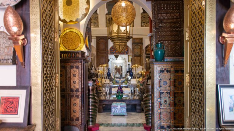 Decorative-arts showroom seen through an ornate doorway, filled with carved furniture, metal lamps, ceramics, mirrors, and framed works.