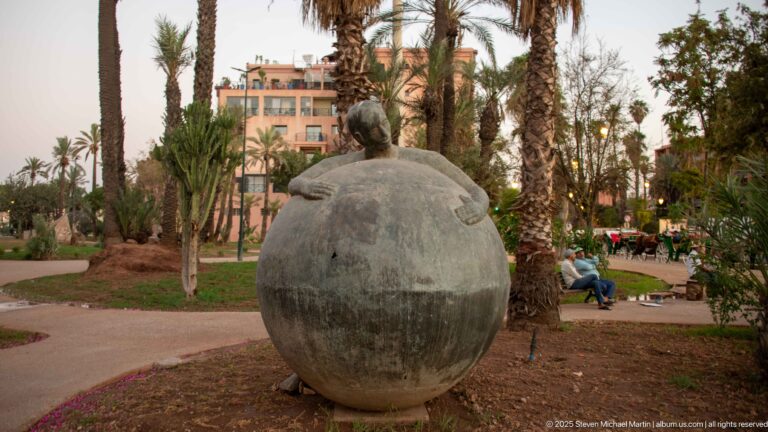 Rounded bronze seated figure viewed from behind in a palm-filled landscaped garden in Marrakech.
