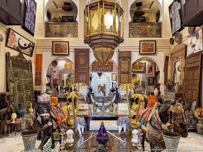 Large Marrakech antiques showroom packed with carved furniture, textiles, ceramics, sculptures, arches, balconies, and a monumental hanging lantern.