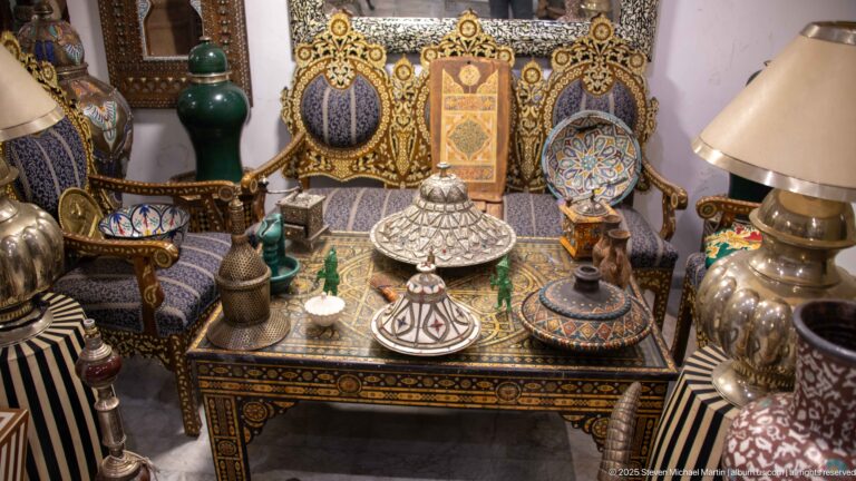 Decorative-arts display with an inlaid table, painted ceramics, metal vessels, lamps, and patterned upholstered furniture.