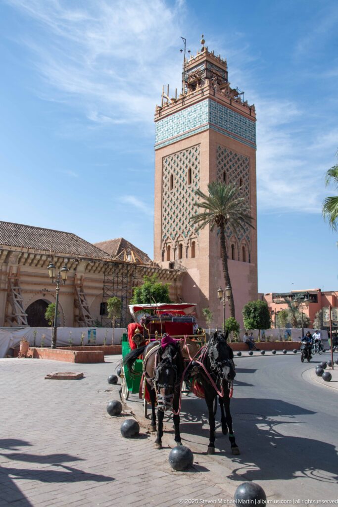 Koutoubia minaret with restoration scaffolding above the mosque complex and a horse-drawn carriage in the foreground.