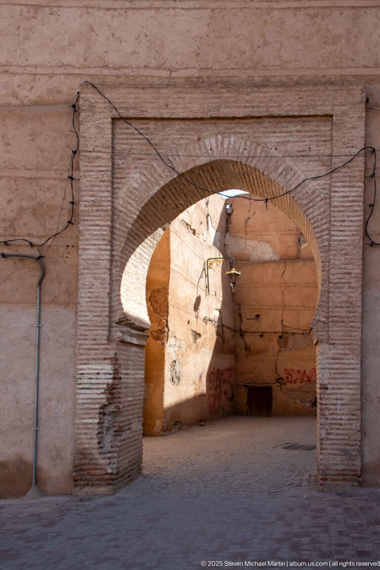 Large pointed horseshoe arch in a weathered pink wall with brick edging, patched masonry, wiring, and a shaded passage beyond.