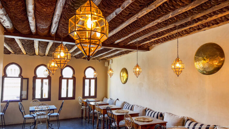 Kasbah Café dining room with timber-and-reed ceiling, hanging metal lanterns, arched windows, tables, and a long cushioned banquette.