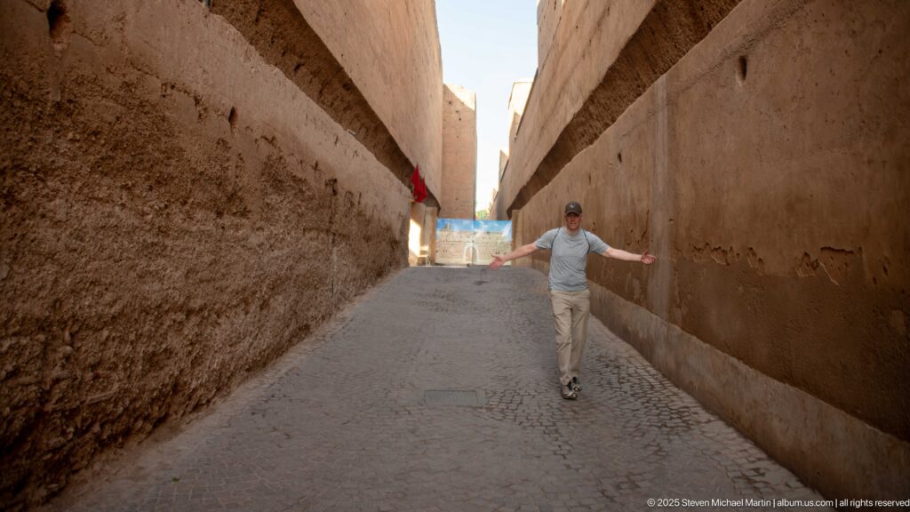 Visitor walking through a narrow paved passage between tall earthen walls inside Badii Palace.