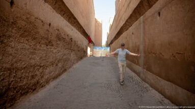 Visitor walking through a narrow paved passage between tall earthen walls inside Badii Palace.