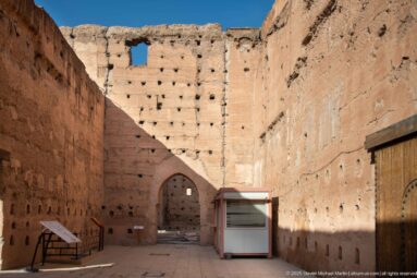 Ruined interior enclosure at Badii Palace with perforated earthen walls, an arched doorway, and modern visitor fixtures.