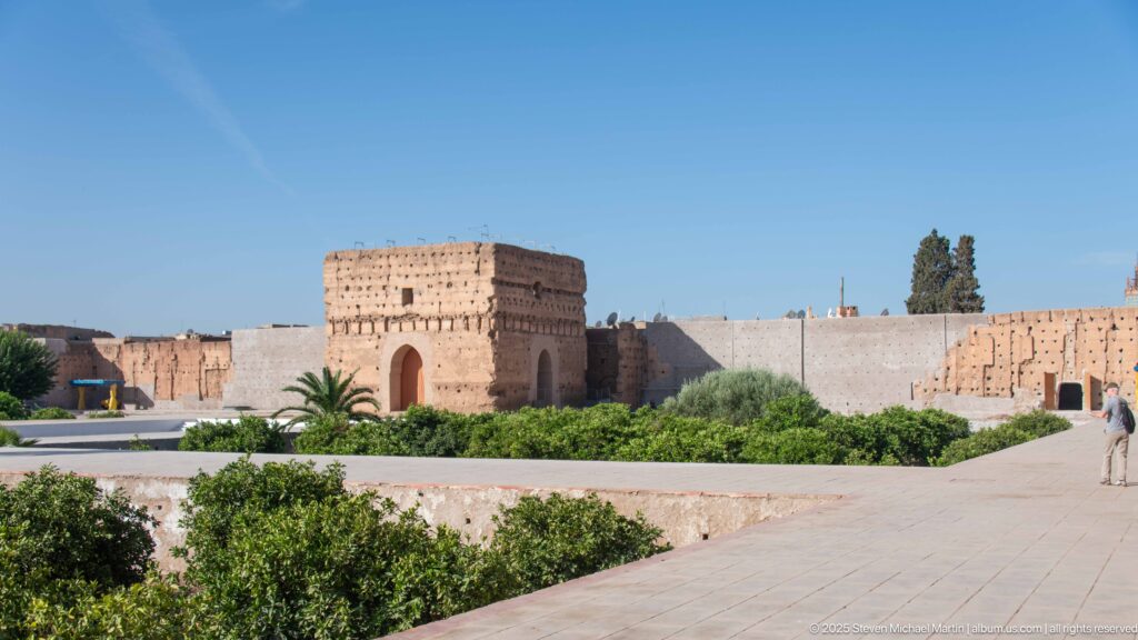 Rectangular earthen pavilion with a tall arched opening beyond gardens and paved paths inside Badii Palace.