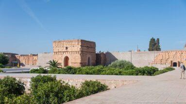 Rectangular earthen pavilion with a tall arched opening beyond gardens and paved paths inside Badii Palace.