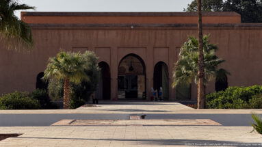 Low plastered building with three arched openings behind palm trees and formal planting inside the Badii Palace complex.