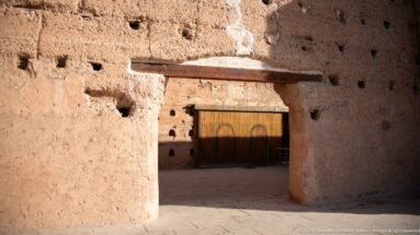 Deep rectangular opening through a thick earthen wall at Badii Palace, framing a closed metal gate beyond.