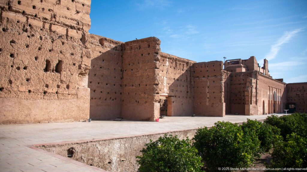 Layered view of massive earthen walls, openings, paved courts, and low greenery inside the Badii Palace ruins.