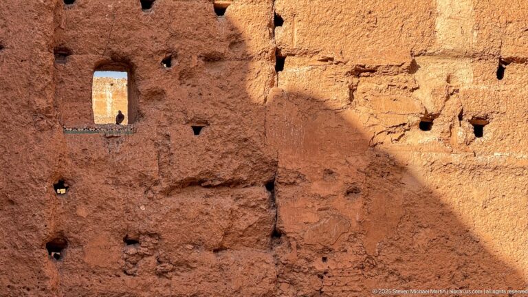 Close detail of eroded reddish earthen masonry at Badii Palace with small holes, a narrow opening, and diagonal shade.