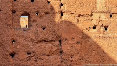 Close detail of eroded reddish earthen masonry at Badii Palace with small holes, a narrow opening, and diagonal shade.