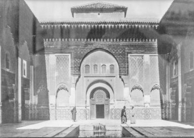 Historic black-and-white view of an ornate Moroccan pavilion with a horseshoe arch, tiled walls, upper gallery, and water channel.
