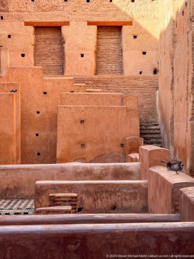 Sunlit Badii Palace interior with low earthen partitions, patterned tile floors, steps, wooden shutters, and a pigeon.