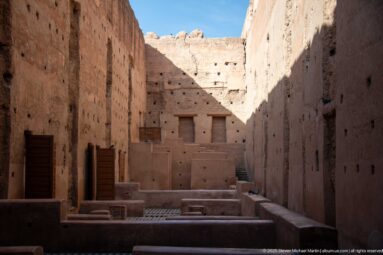 Long shadowed interior at Badii Palace with low partitions, geometric tile floors, timber shutters, and bright sunlight above.