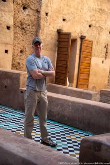 Visitor standing with arms crossed on geometric tile flooring between low earthen walls inside Badii Palace.