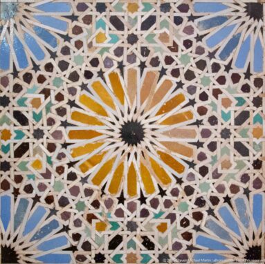 Close-up of a multicolored zellij rosette with ochre radial petals, interlaced white star patterns, and blue-green accents.