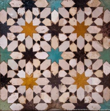 Close-up of worn zellij with ochre and turquoise eight-pointed stars among white, black, and green geometric tiles.