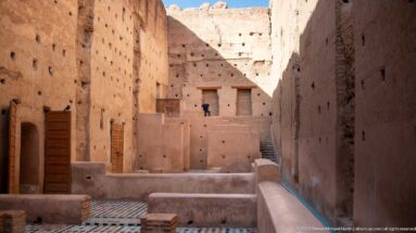 Elevated view through Badii Palace interior bays with geometric tile floors, timber shutters, steps, and a distant visitor.