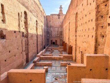 Axial elevated view down a long Badii Palace interior with repeated low walls, zellij floors, wooden shutters, and a distant doorway.