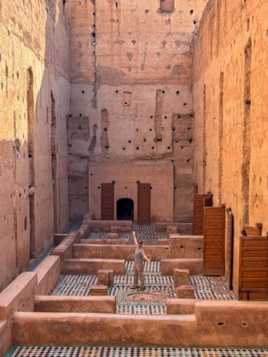 Visitor walking among low partitions and geometric tile floors beneath high earthen walls at Badii Palace.