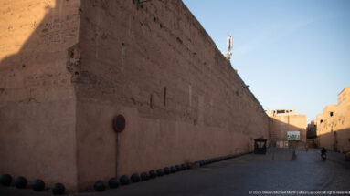Massive earthen wall of Badii Palace lining a shaded lane, with dark bollards leading toward a sunlit opening.