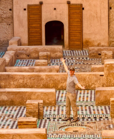 Visitor posing with arms raised over a circular basin and geometric zellij floor inside Badii Palace.