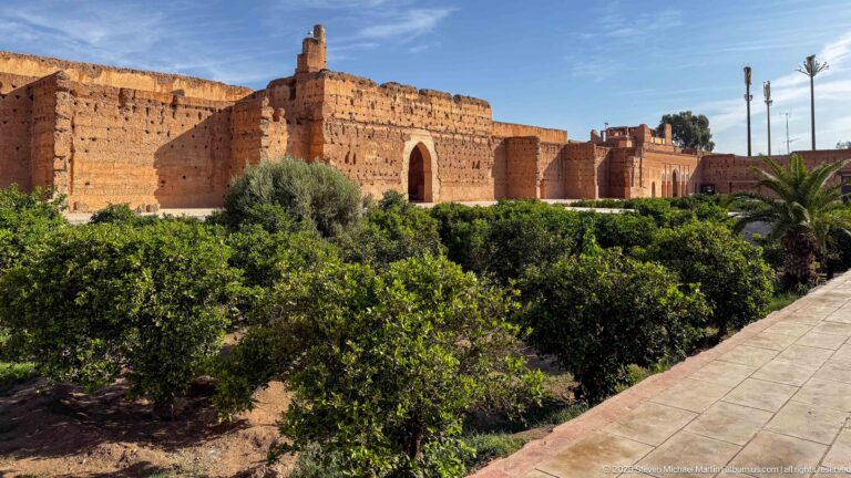 Wide view of Badii Palace walls behind dense green garden trees and palms, with a paved visitor path at right.