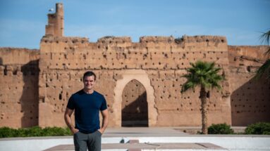 Visitor standing before a tall pointed arch in a weathered earthen wall with a palm tree at Badii Palace.