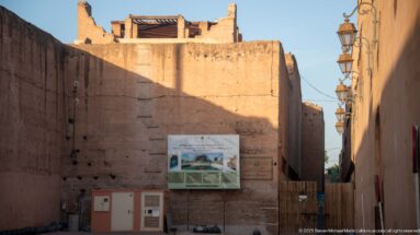 Narrow approach to Badii Palace between high earthen walls, with interpretive signage and decorative lamps in bright sunlight.